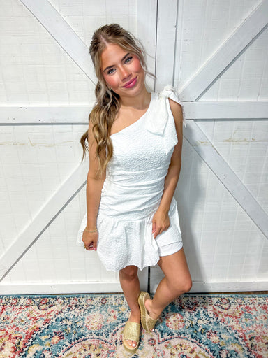 Woman modeling a white shoulder mini dress with a white bow at the shoulder. The material is ruched in the middle around the waist and the dress has a white textured pattern all over.