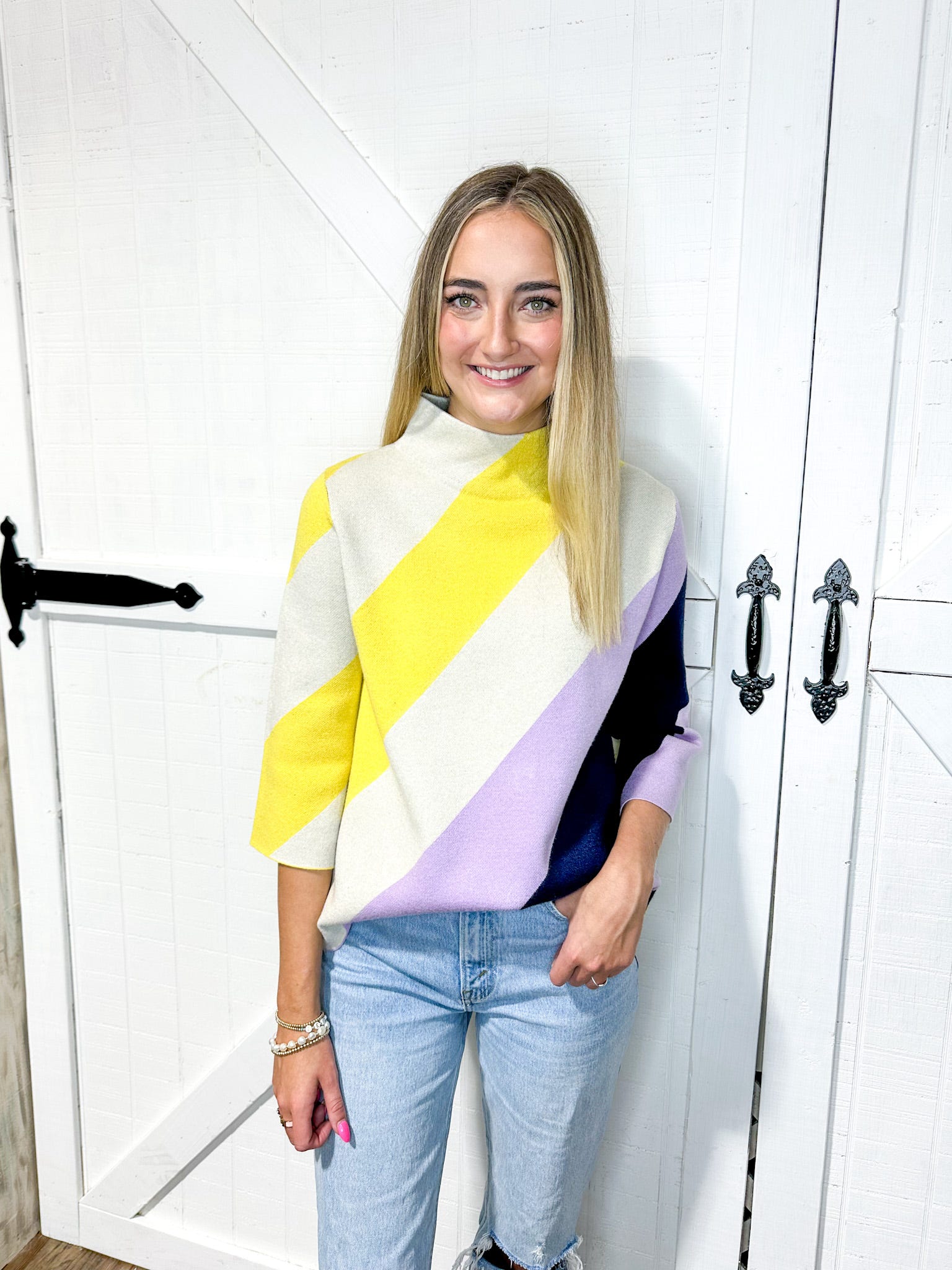 Woman modeling a mock-neck sweater with three quarter sleeves that has thick diagonal stripes in cream, yellow, lavender and navy