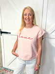 Woman modeling a light pink short sleeve sweatshirt that says love in red cursive letters. The sweatshirt has a texture pattern of small hearts all over.
