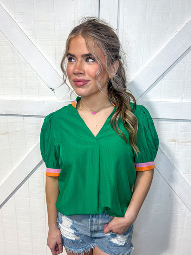 Woman modeling a green v-neck blouse with an orange and pink stripe around the neck and sleeve edge