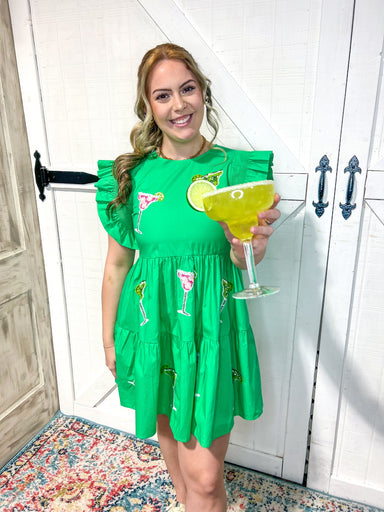 Another view of a Woman modeling a kelly green tiered mini dress with sequined margarita glasses all over. Model is also holding a fake margarita glass in her hand. The dress has a cap ruffle sleeve