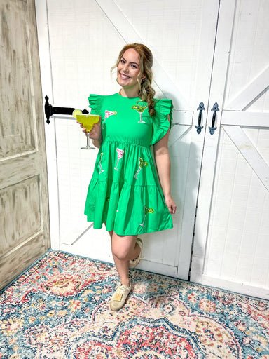 Woman modeling a kelly green tiered mini dress with sequined margarita glasses all over. Model is also holding a fake margarita glass in her hand. The dress has a cap ruffle sleeve