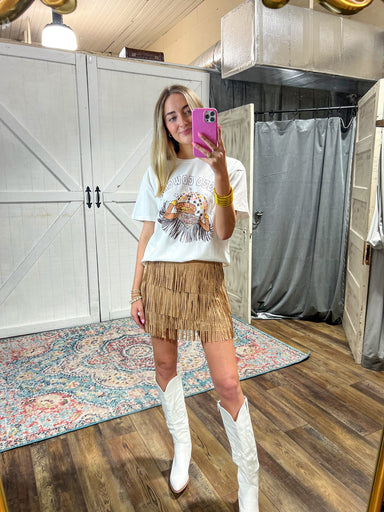 Model taking a selfie in front of a full length mirror wearing a white short sleeve tee saying Disco Cowgirl and showing a graphic disco ball and the back of a female's jacket with fringe. All in colors of brown, light pink and yellow
