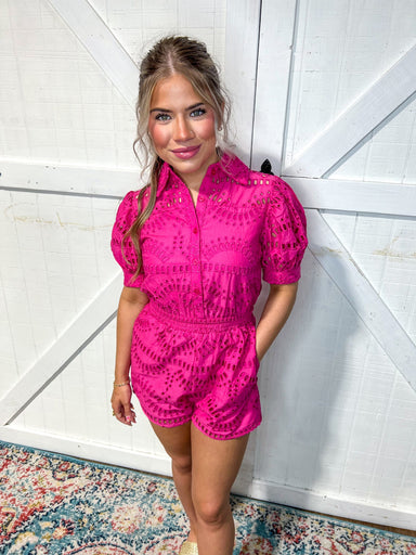 Woman modeling a hot pink/ magenta short sleeve romper with a collar, buttons, cinch waist and attached shorts 