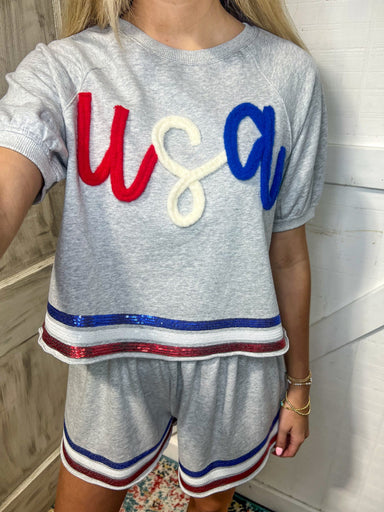 Up close photo of grey cotton shorts set with red, white and blue sequins trim around sleeves, bottom of shirt and bottom of shorts saying USA in red, white and blue yarn