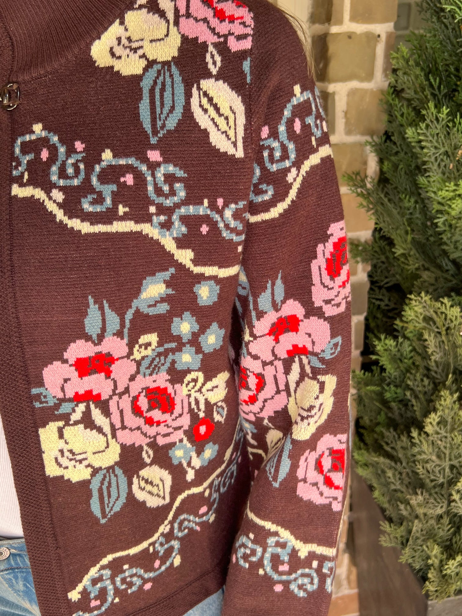 Close-up of the sleeve of a brown cardigan with floral patterns in front of a brick wall.