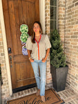 Woman wearing a cream and khaki striped short sleeve blouse with an orange and blue stripe around the collar and down the front standing outside a house with a wooden door and brick wall.