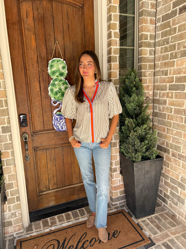 Woman wearing a cream and khaki striped short sleeve blouse with an orange and blue stripe around the collar and down the front standing outside a house with a wooden door and brick wall.