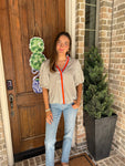Woman wearing a cream and khaki striped short sleeve blouse with an orange and blue stripe around the collar and down the front standing outside a house with a wooden door and brick wall.