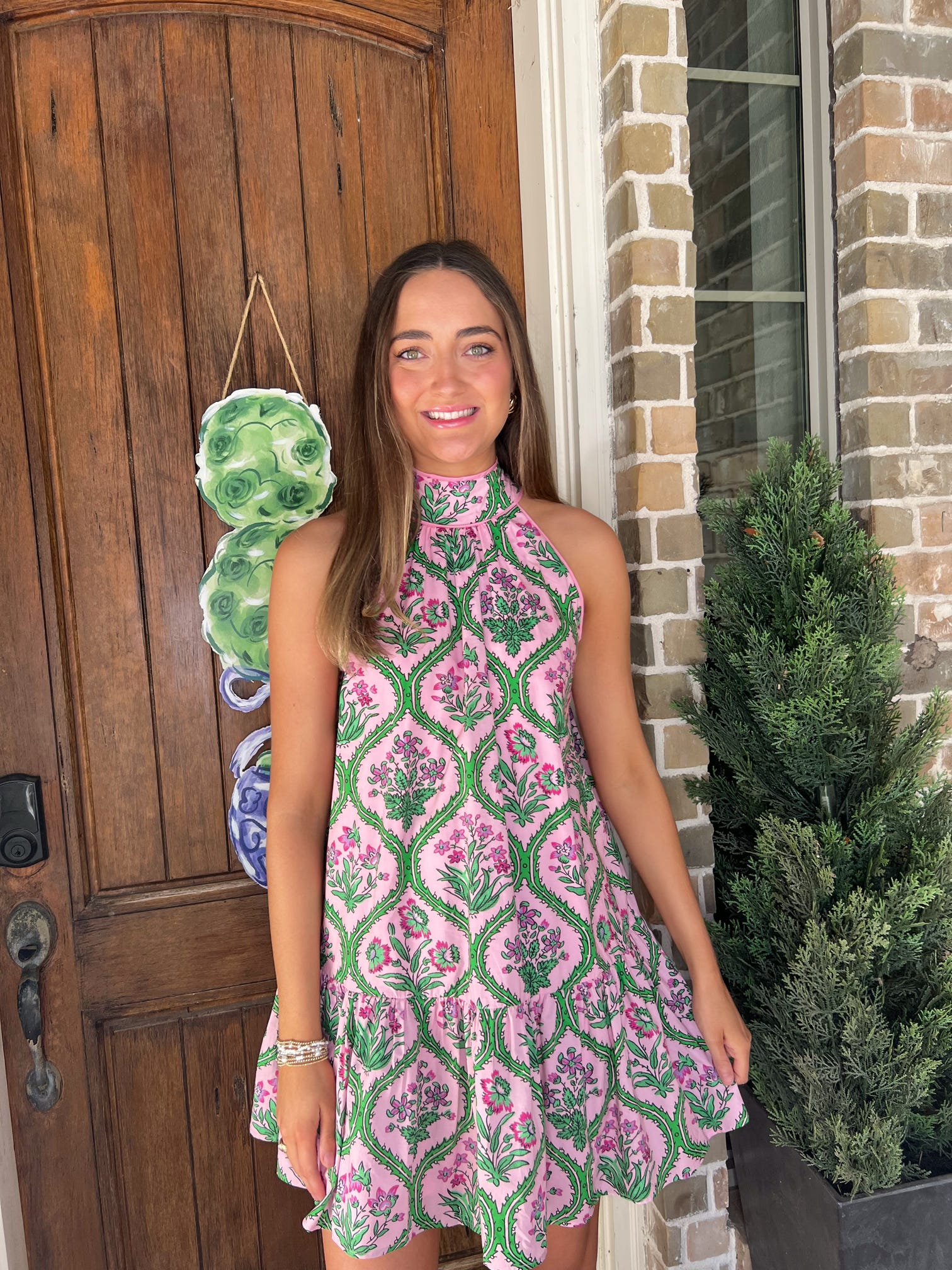 Woman in a pink and green patterned dress standing in front of a wooden door.