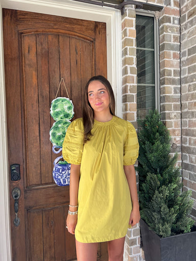 Woman in a chartruse colored dress standing outside a house with a wooden door and brick wall.