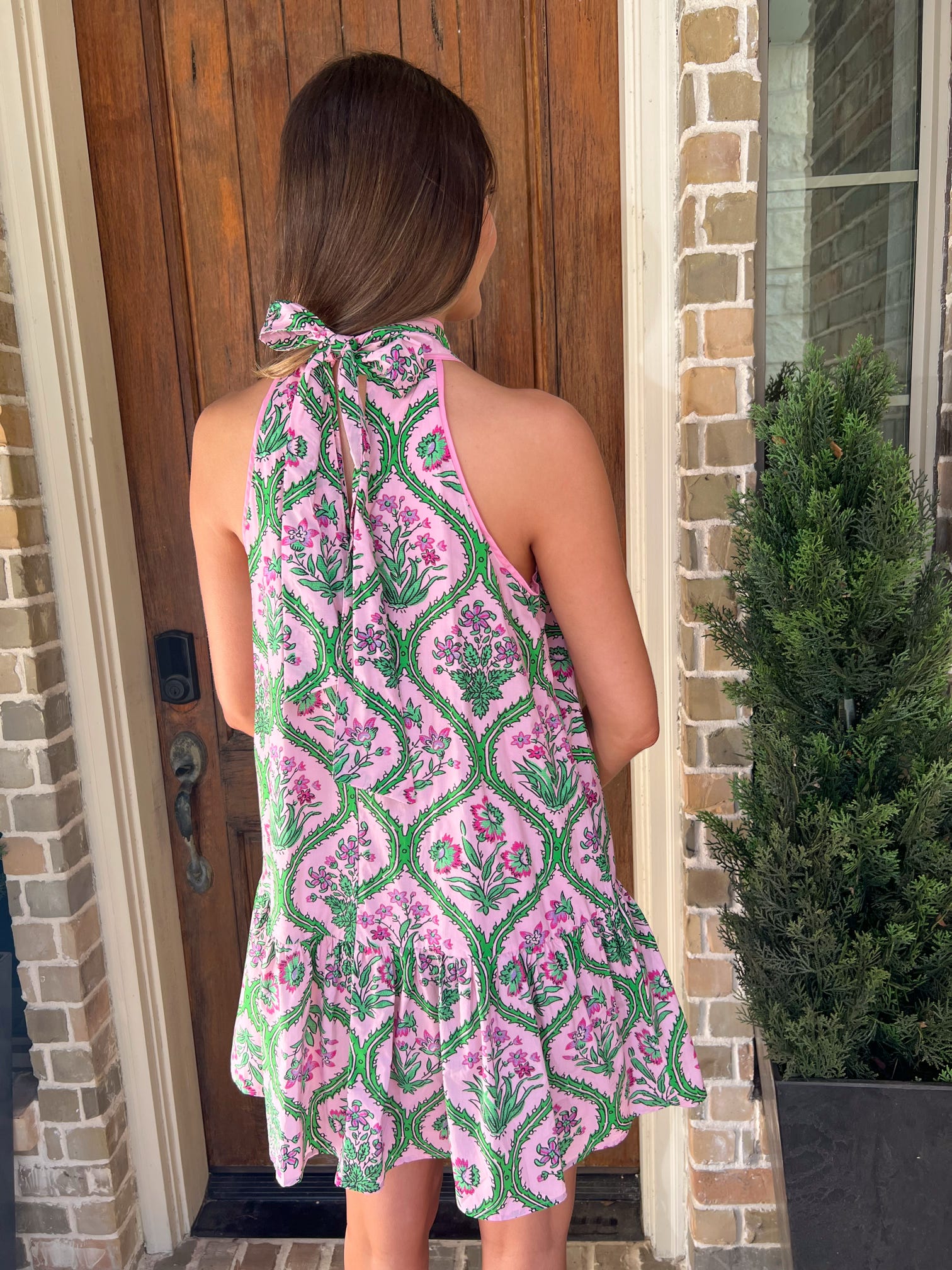 Photo of the back of the dress of a woman wearing a pink and green patterned dress standing in front of a brick house.