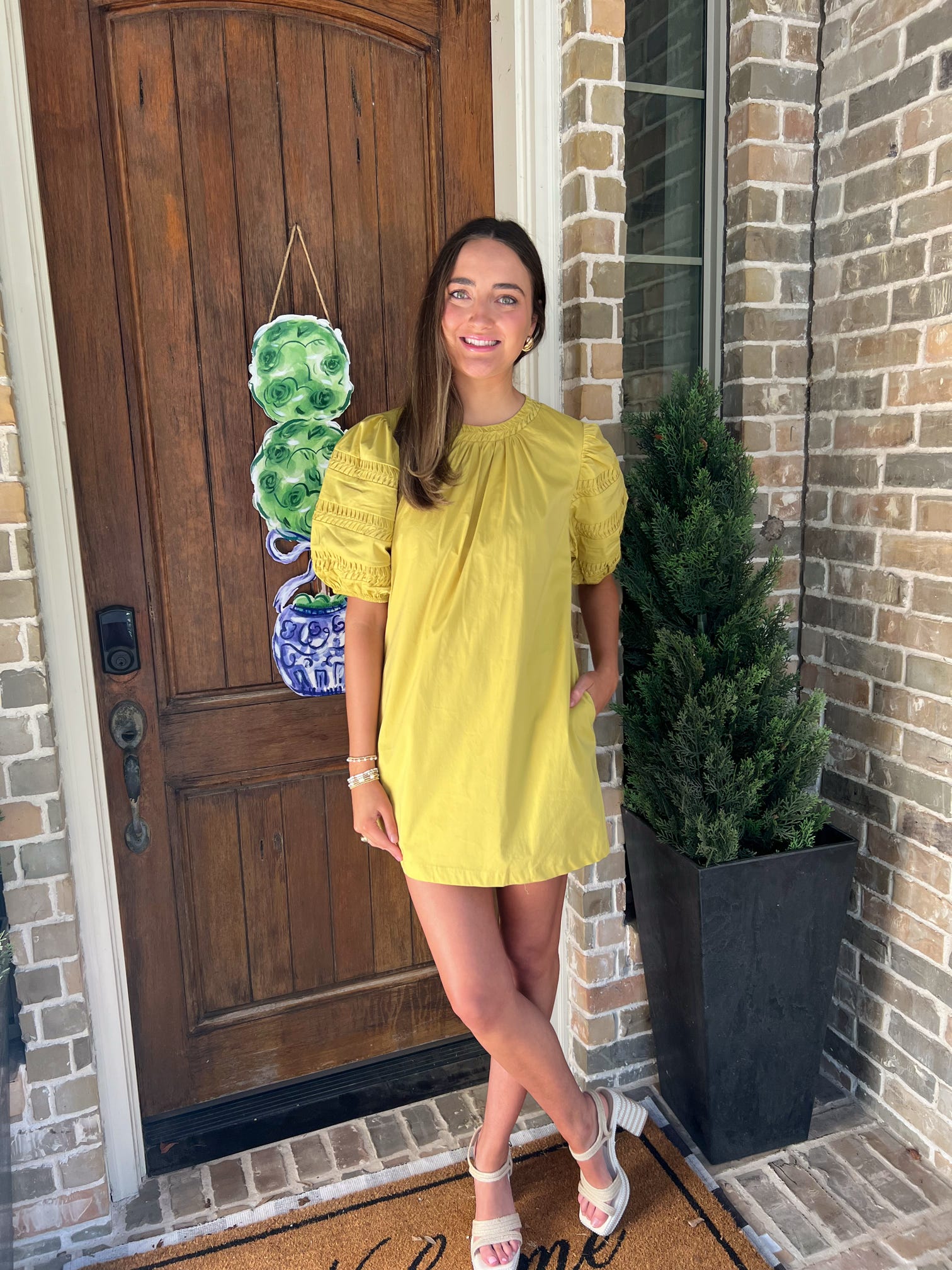 Woman in a chartruse colored dress standing outside a house with a wooden door and brick wall.