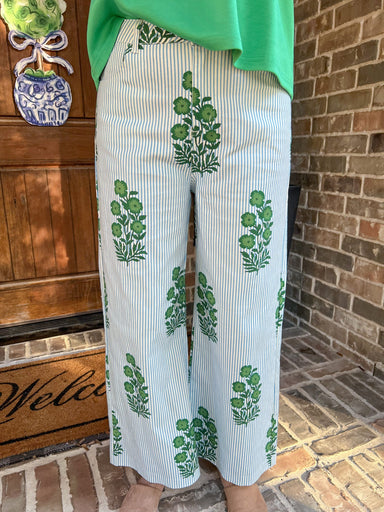 Up close photo of a woman wearing white pants with green floral patterns in front of a brick wall.