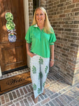 Woman in green shirt and light blue pants with floral patterns standing in front of a brick wall.