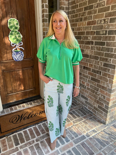 Woman in green shirt and light blue pants with floral patterns standing in front of a brick wall.