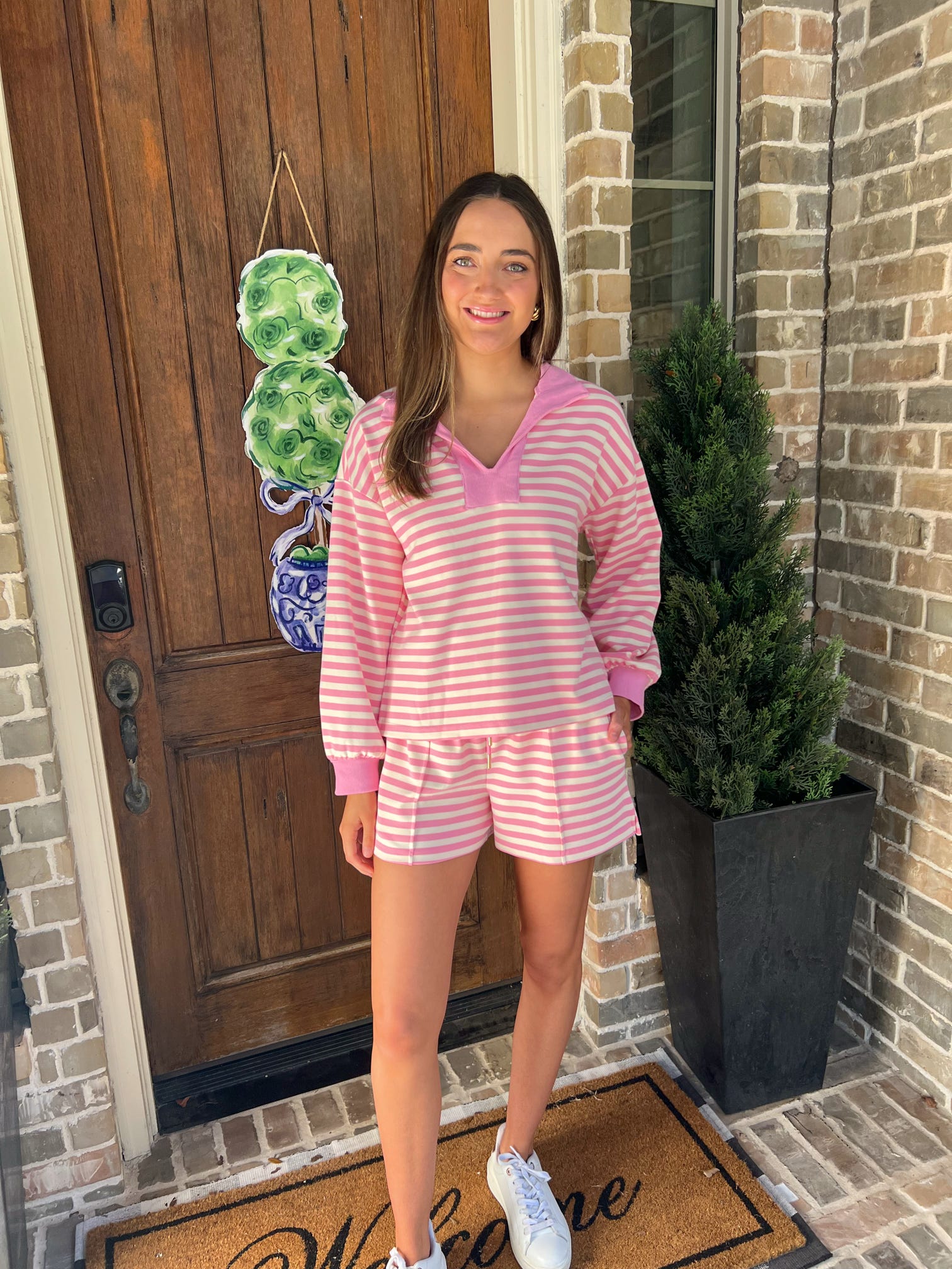Woman in pink striped shorts set standing outside a house with a welcome mat.