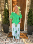 Woman in green shirt and white pants with green patterns standing on a porch.