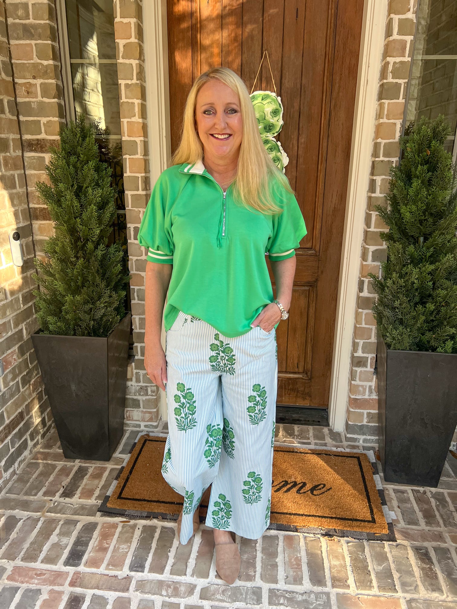 Woman in green shirt and white pants with green patterns standing on a porch.