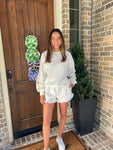 Woman in a cream with khaki polka dots matching long sleeve and shorts set standing in front of a brick house with a wooden door.
