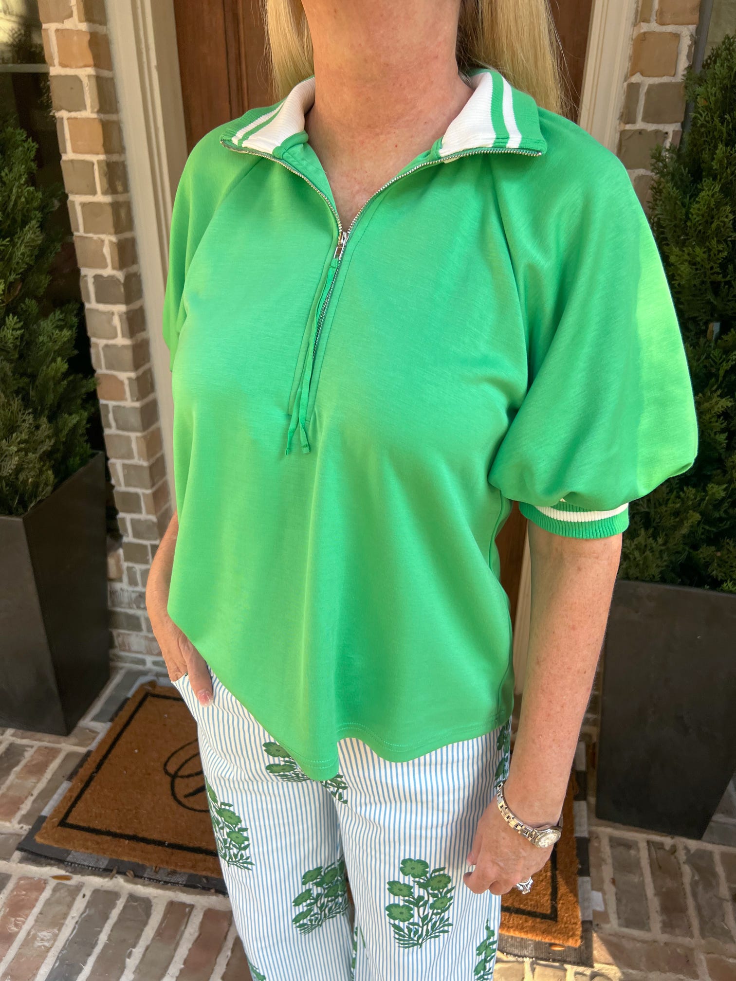 Person wearing a green quarter-zip shirt with white trim in front of a brick wall.