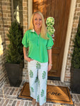 Woman in green shirt and white pants with green patterns standing in front of a door.
