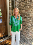 Woman in a green embroidered blouse standing outside a house with a brick wall.