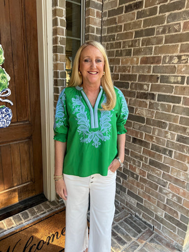Woman in a green embroidered blouse standing outside a house with a brick wall.