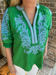 Green blouse with white embroidery worn by a person, standing against a brick wall.