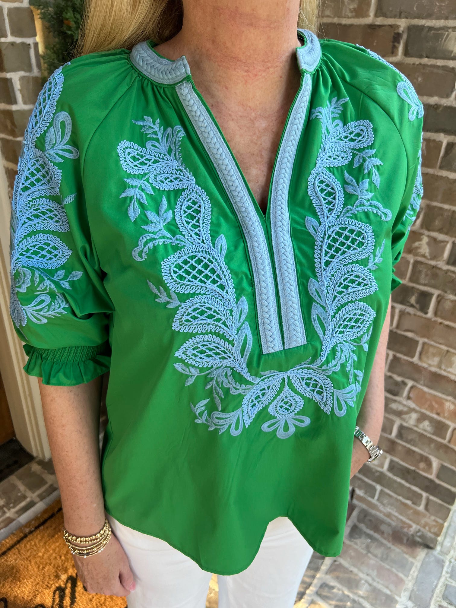 Green blouse with white embroidery worn by a person, standing against a brick wall.