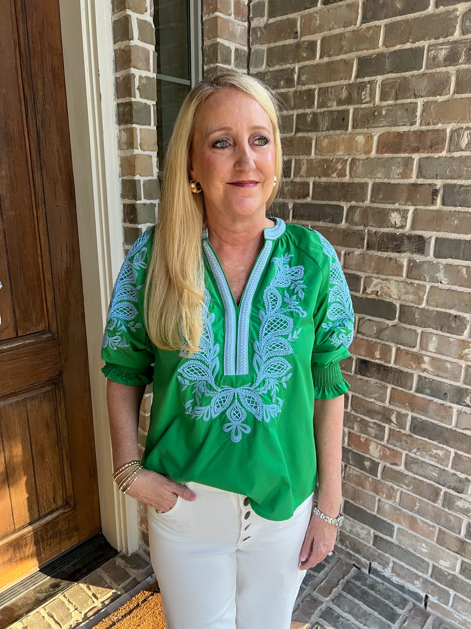Woman wearing a green embroidered top standing in front of a brick wall.