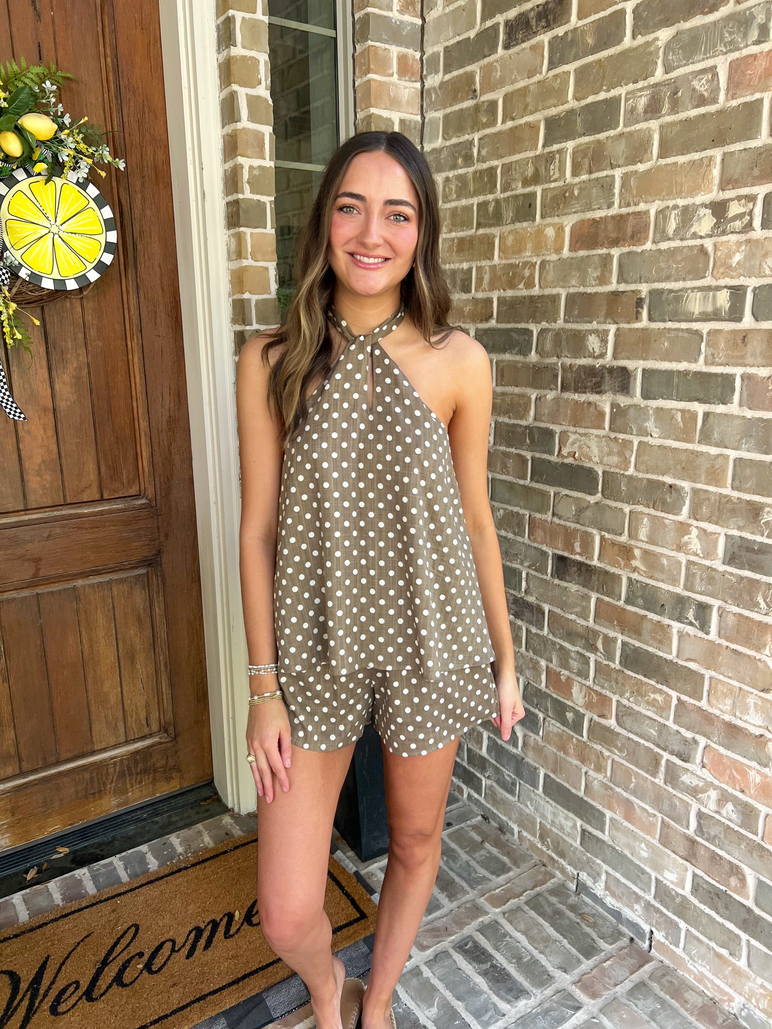 Woman in a polka dot romper standing in front of a brick wall and wooden door.