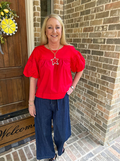 Woman in a red top and blue jeans standing in front of a brick wall.