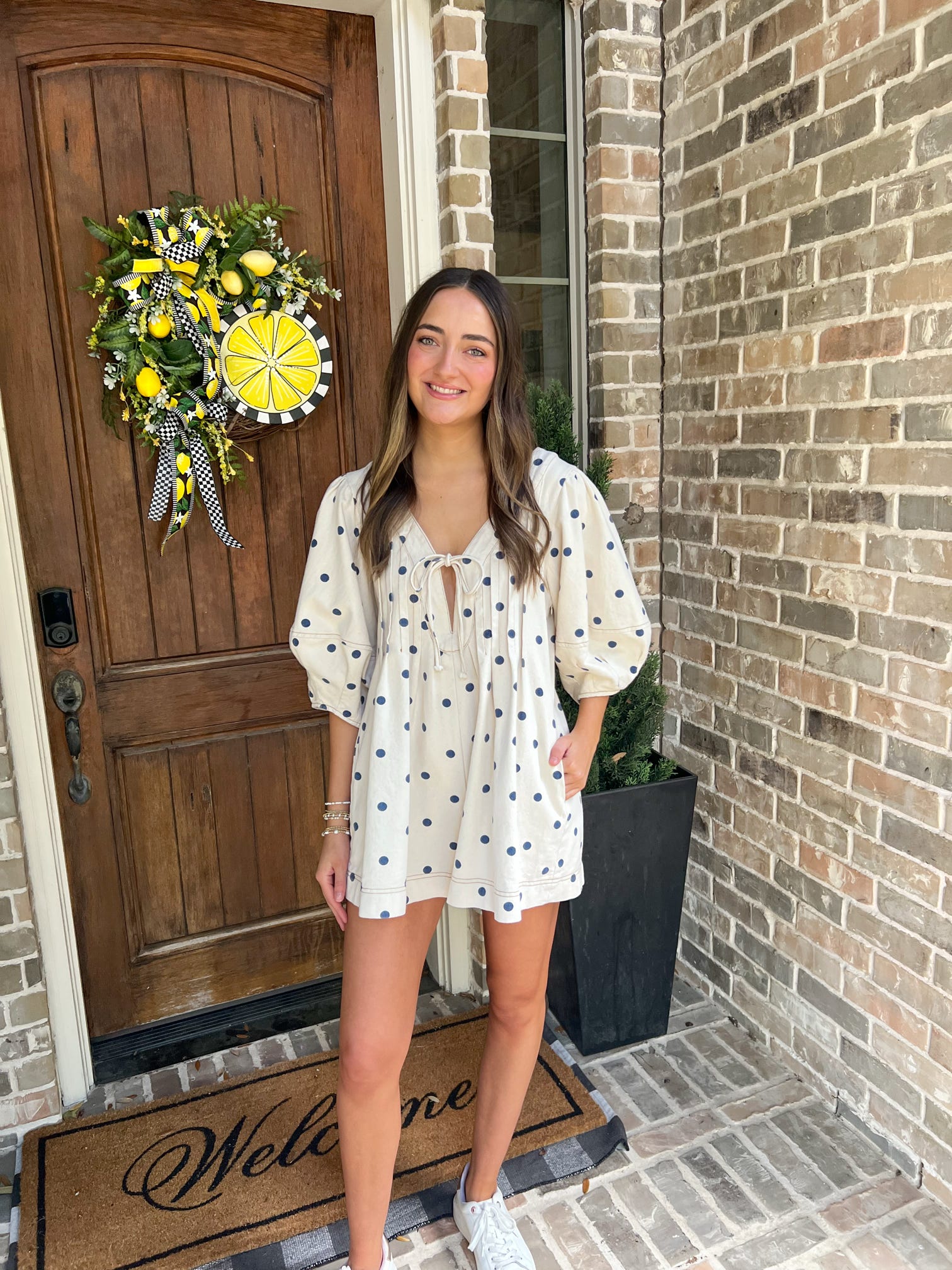 Woman standing in front of a door with a decorative wreath, wearing a white polka dot dress.
