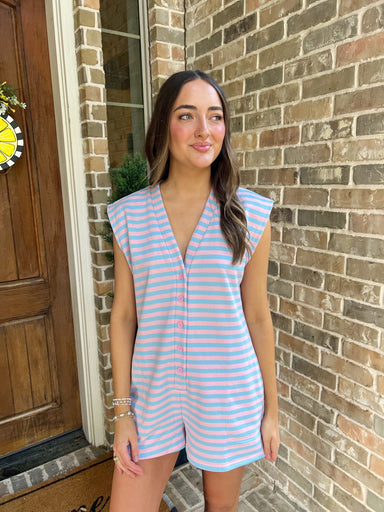 Woman wearing a light pink & baby blue striped sleeveless romper standing in front of a brick wall.