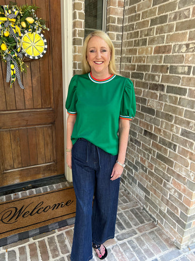 Woman in a green shirt and blue jeans standing in front of a brick house with a wooden door.