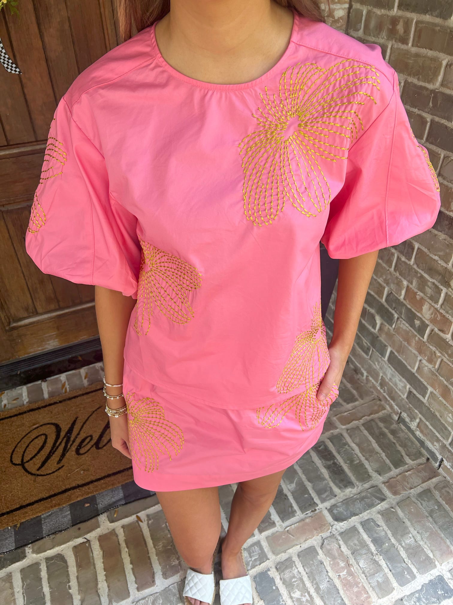 Person wearing a matching pink top & skirt set with gold embroidery standing on a brick porch.