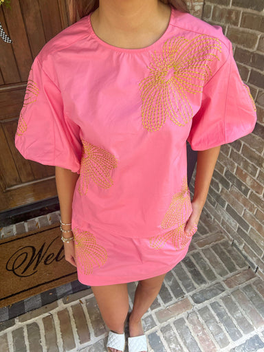 Person wearing a matching pink top & skirt set with gold embroidery standing on a brick porch.