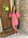 Woman in a pink outfit standing on a porch with a 'Welcome' mat and decorative door wreath.