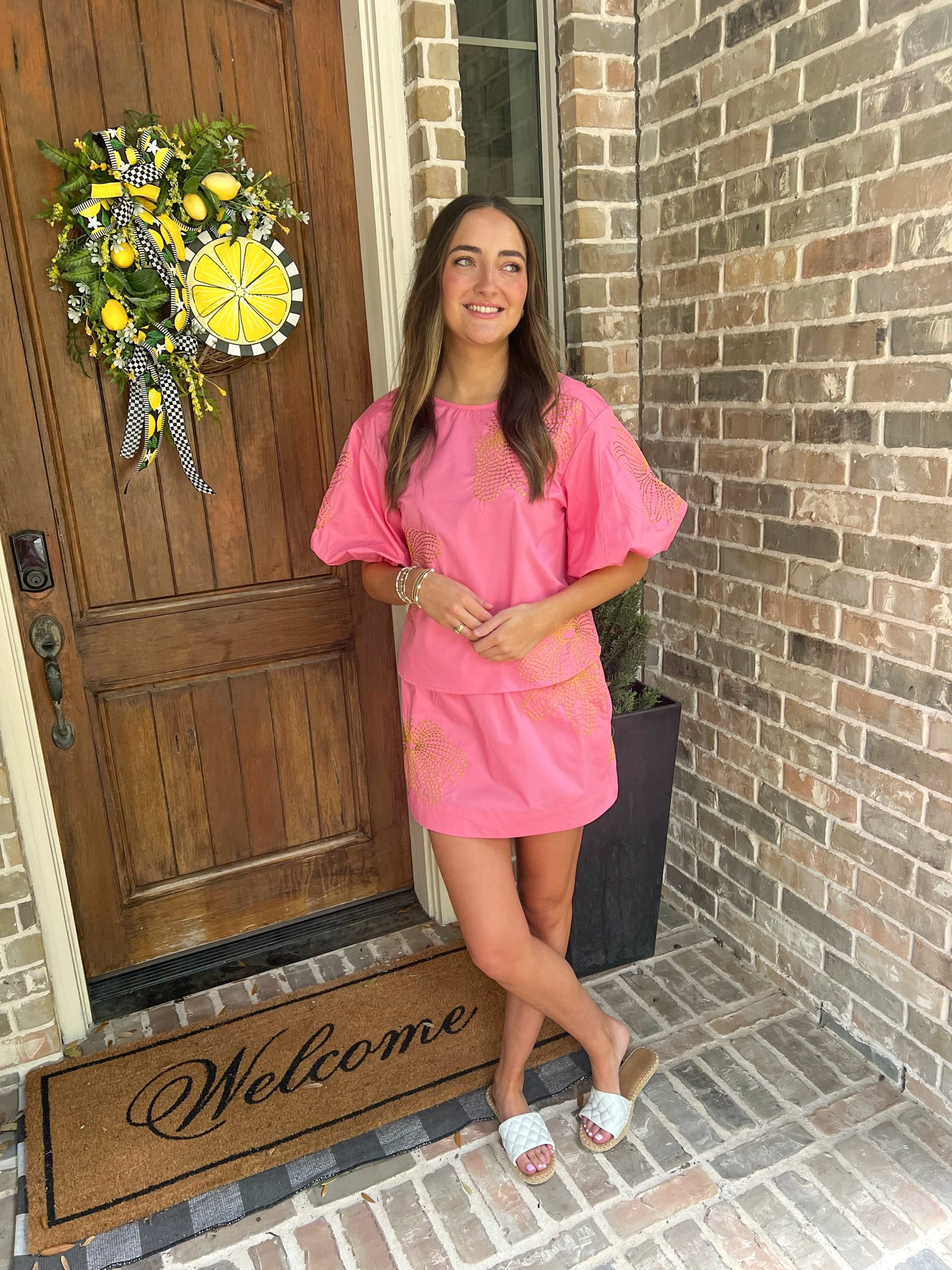 Woman in a pink outfit standing on a porch with a 'Welcome' mat and decorative door wreath.