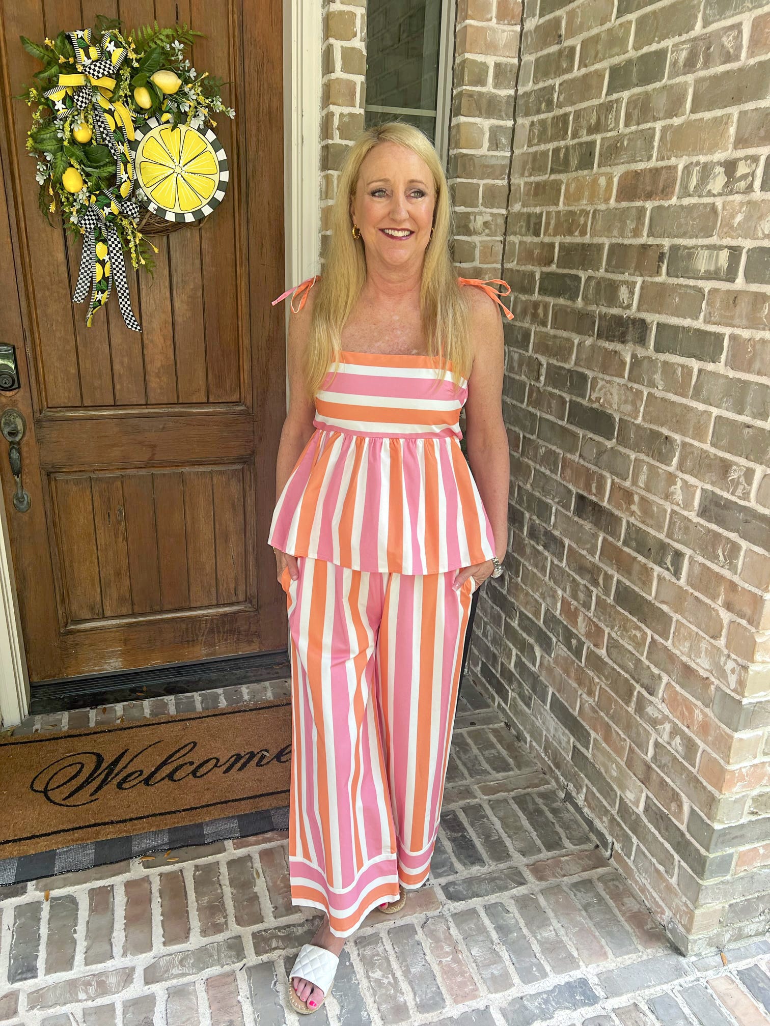 Woman in a striped outfit standing on a porch with a wreath and 'Welcome' mat.