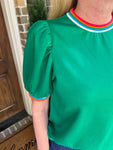Green blouse with rainbow trim worn by a person, standing in front of a door.