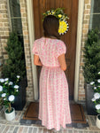 Person wearing a pink floral matching top & skirt set standing in front of a door with a wreath, surrounded by potted plants.