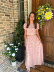 Woman in a pink matching top & skirt set standing in front of a door with a wreath and plants.