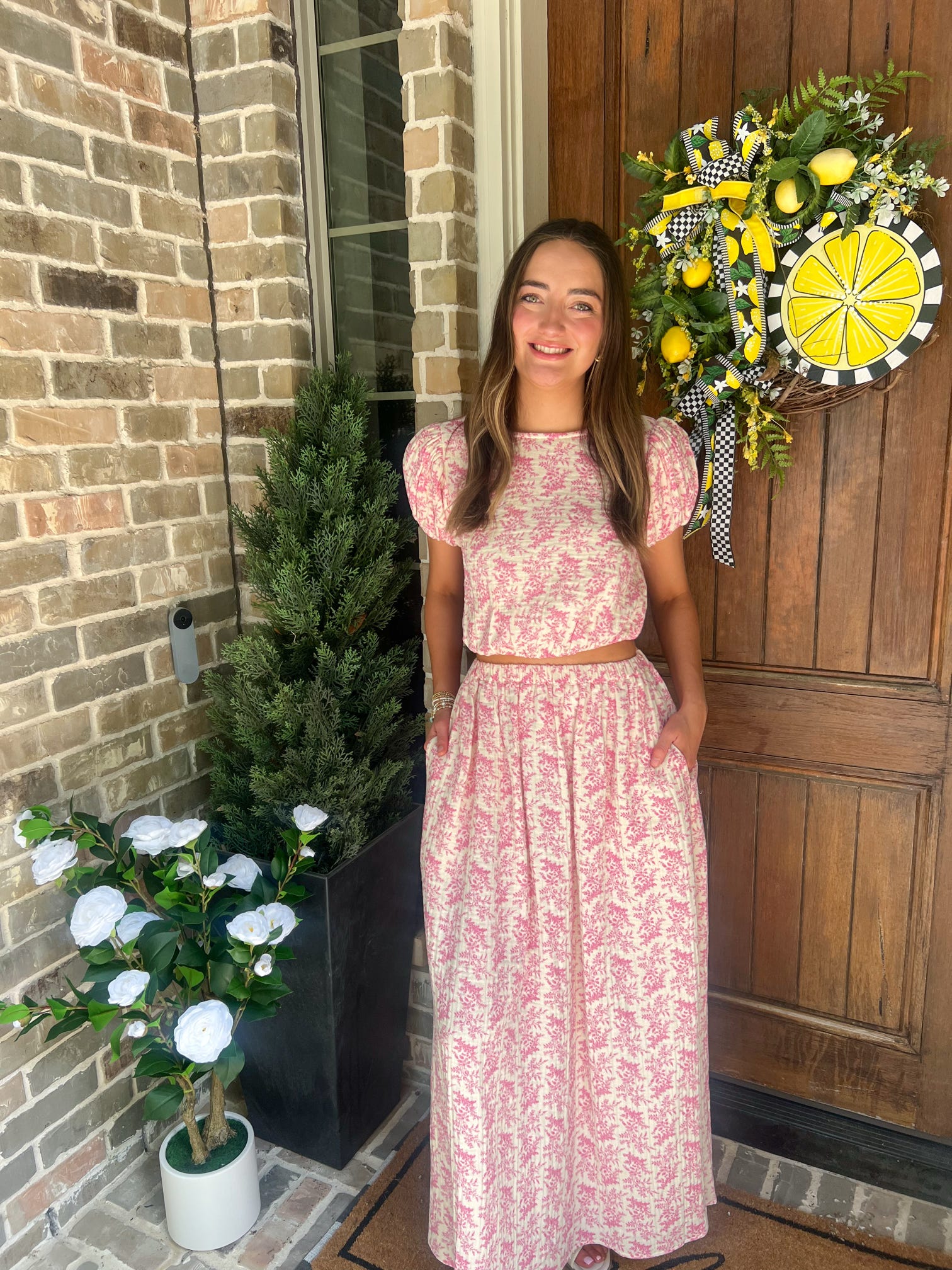 Woman in a pink floral matching top & skirt set standing in front of a door with a wreath, next to potted plants.