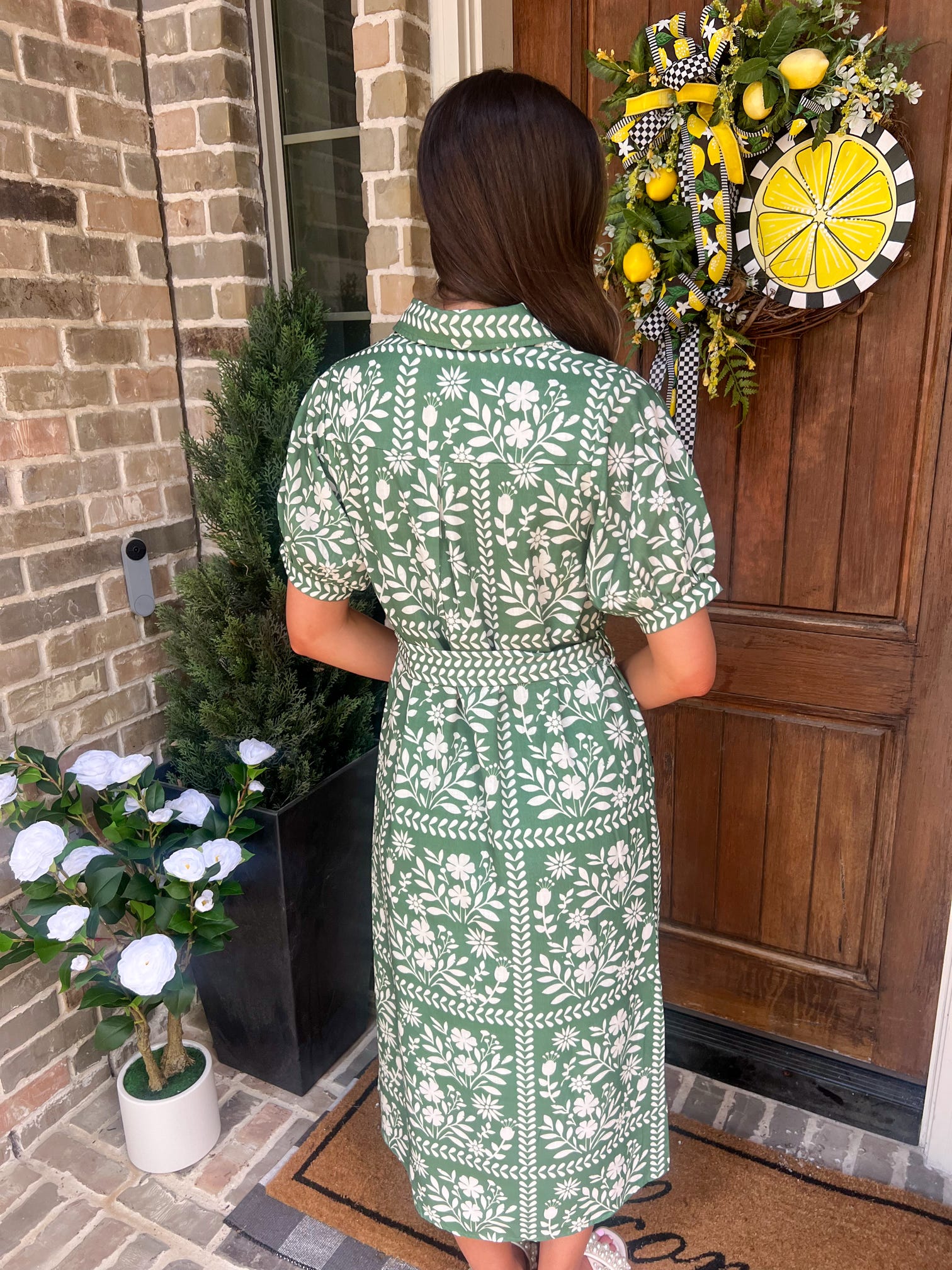 Woman showing the back of a green floral dress standing in front of a door with a decorative wreath.
