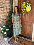 Woman in a green dress standing in front of a door with a decorative wreath and plants.