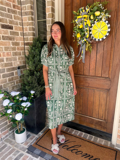Woman in a green dress standing in front of a door with a decorative wreath and plants.