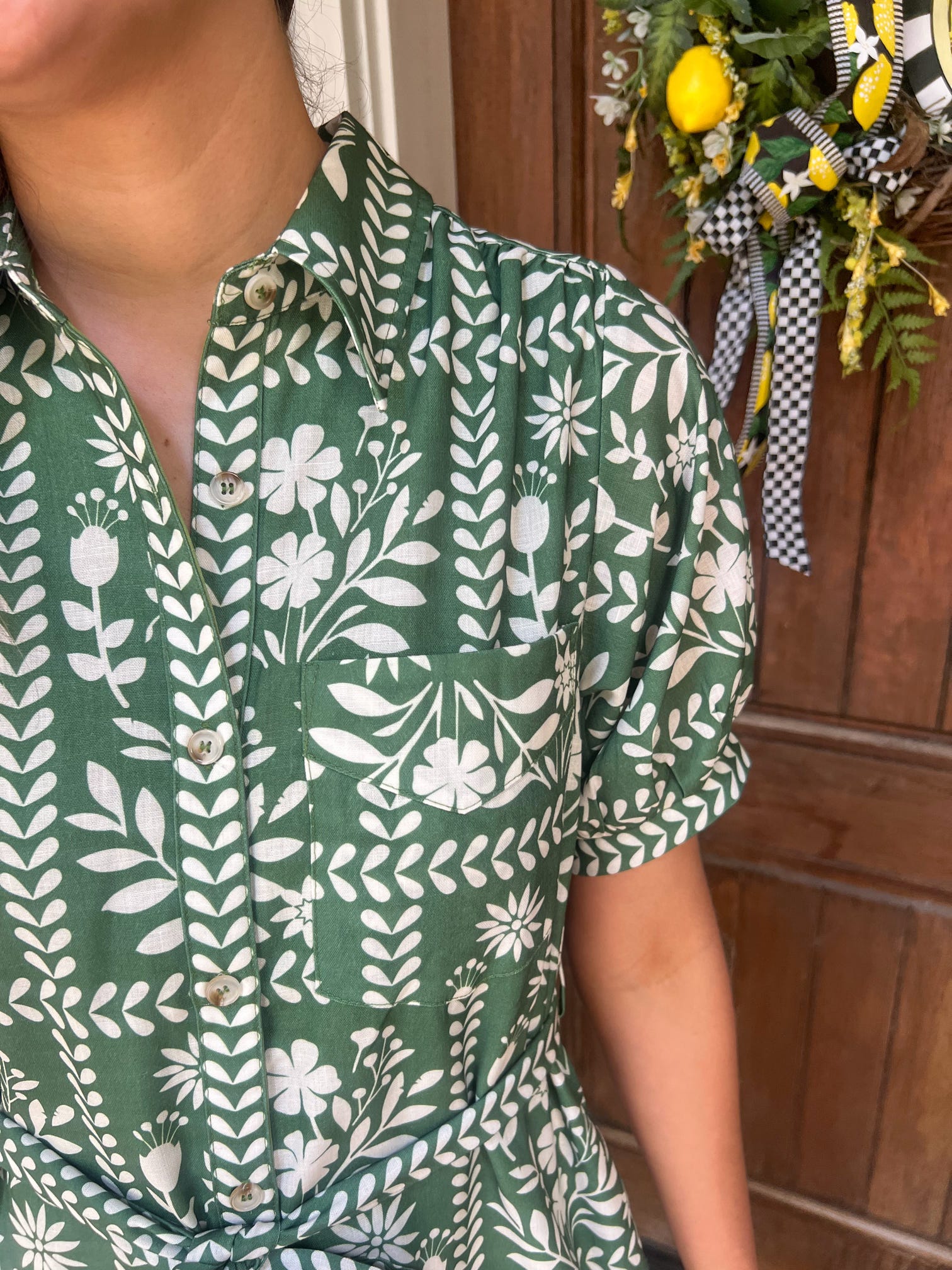 Up close photo of a woman wearing a green floral patterned dress with a wooden door and decorative wreath in the background.