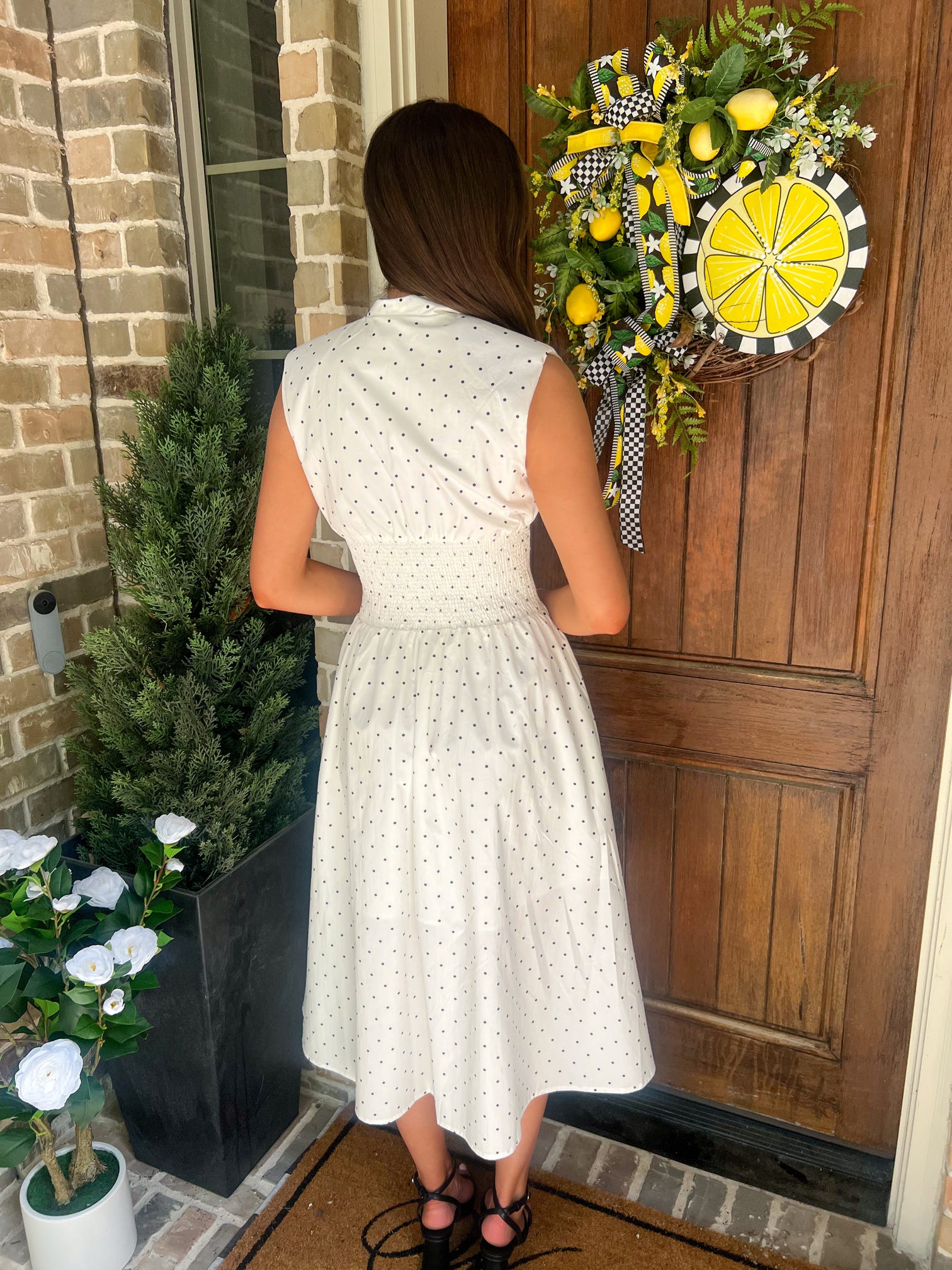 Woman in a white with small black polka dots dress standing in front of a wooden door with a decorative wreath.